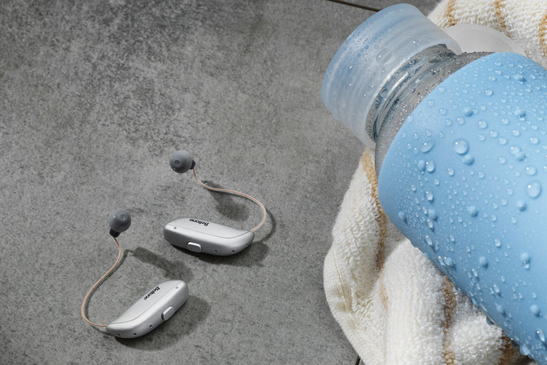 Audifonos Achieve modelo RIE color gris en el piso junto a botella cubierta con gotas de agua sobre una toalla amarillo claro