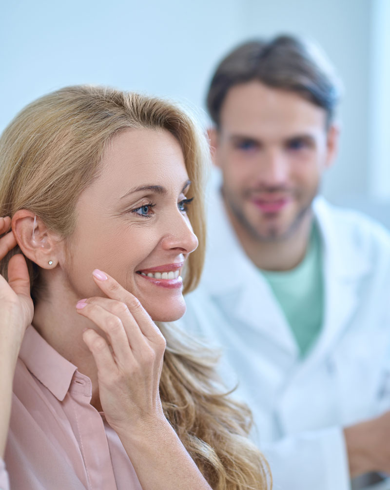 Paciente adulta adaptandose audífono rie en su oreja derecha y a su lado el audioprotesista con bata blanca