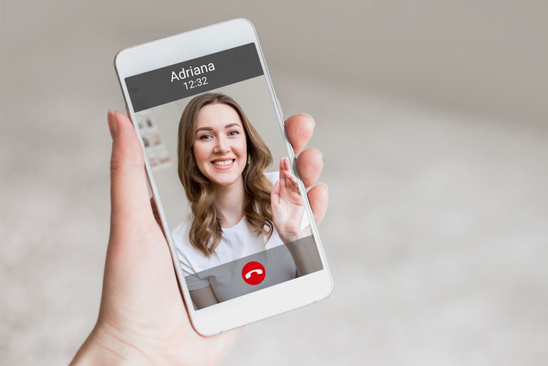 una mano izquierda femenina sosteniendo teléfono movil en video llamada con imagen de mujer joven de nombre Adriana